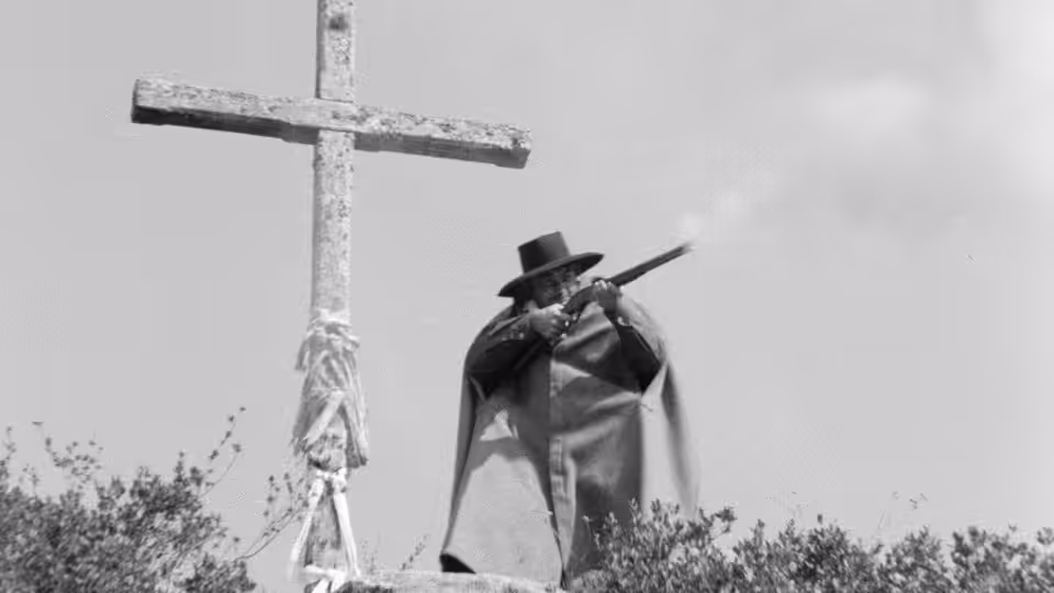 A gunman in a broad hat stands beside a wooden cross above the scrub