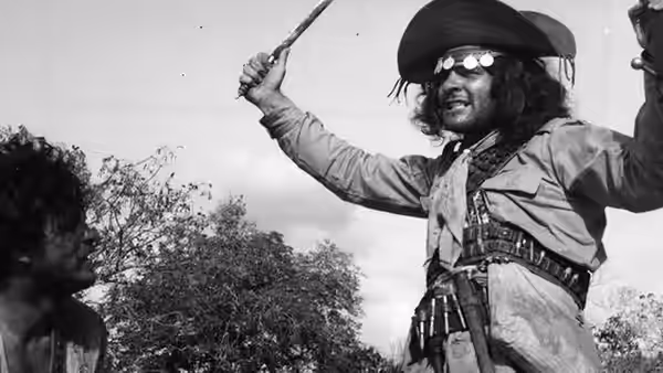 Corisco (Othon Bastos) raises a weapon beside another man in the scrub