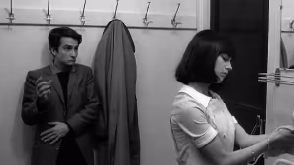 Paul (Jean-Pierre Léaud) stands beside a coat rack as Madeleine Zimmer (Chantal Goya) operates equipment