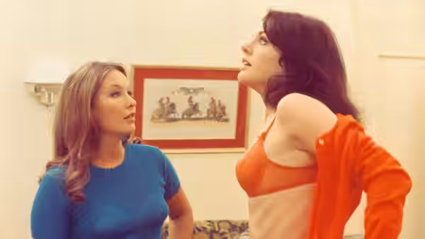 A woman in an orange top stands beside a blonde woman indoors