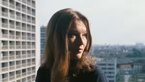 A dark-haired woman looks across the city from a high-rise window