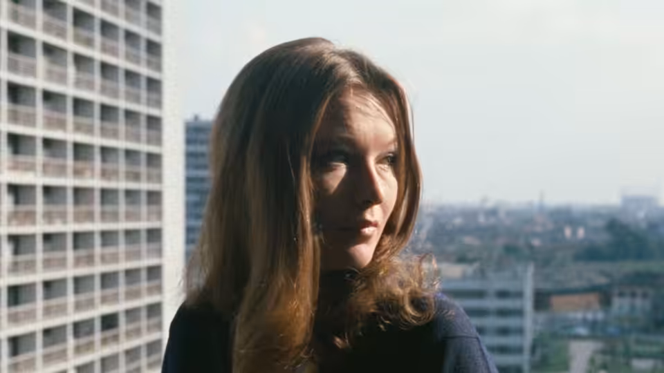 A dark-haired woman looks across the city from a high-rise window