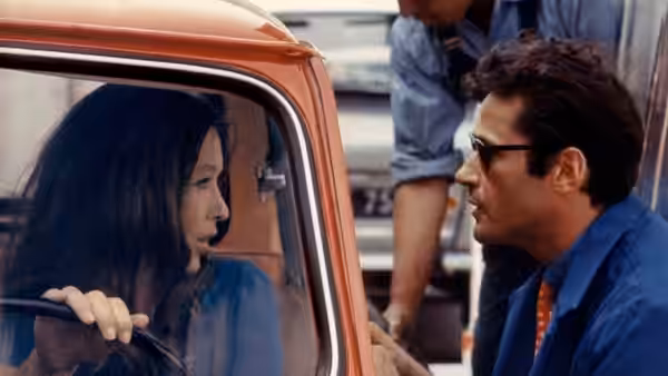 A dark-haired woman sits in a car facing a man in sunglasses
