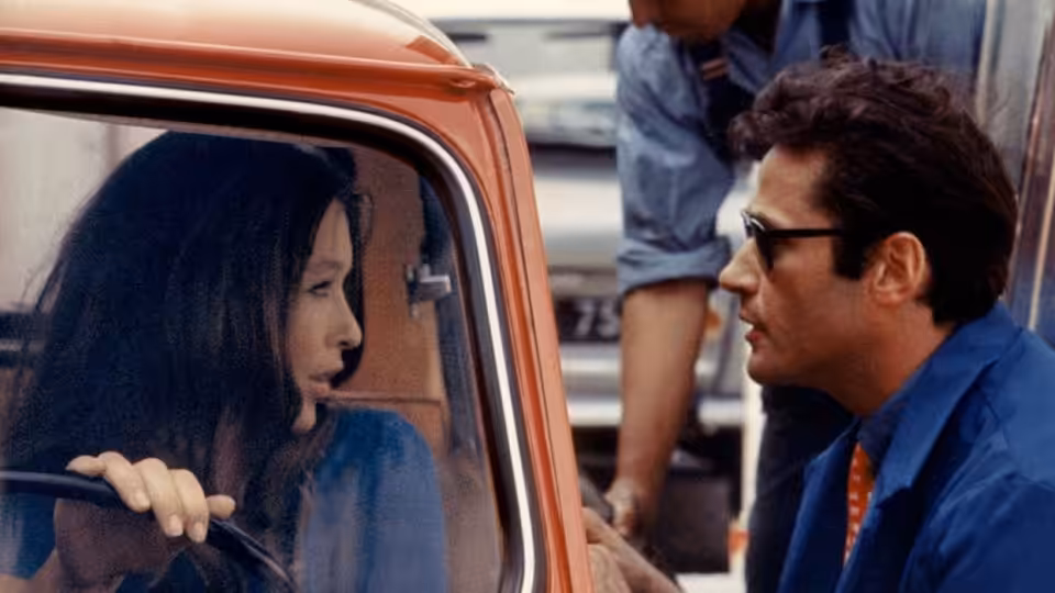A dark-haired woman sits in a car facing a man in sunglasses