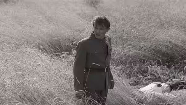A uniformed soldier stands in tall grass beside a body lying on the ground
