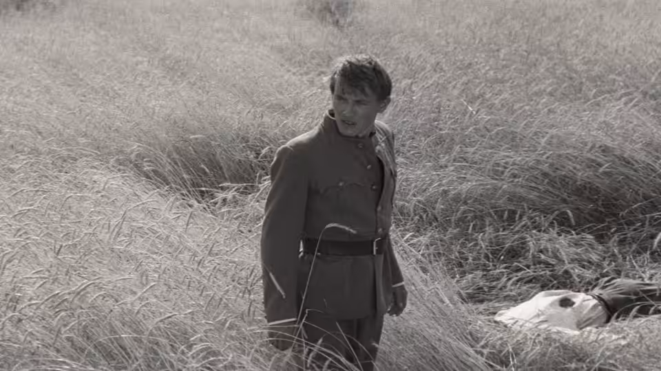 A uniformed soldier stands in tall grass beside a body lying on the ground