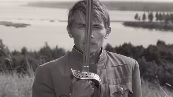 A young uniformed soldier holds a rifle upright before a riverside field