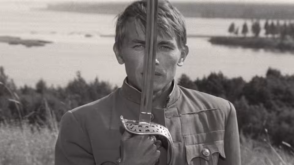 A young uniformed soldier holds a rifle upright before a riverside field