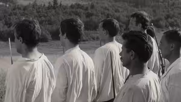 Young men in white shirts stand in a row facing across open ground