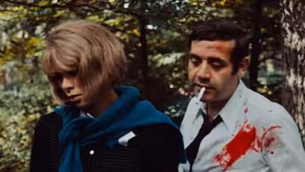 Corinne walks through the woods beside Roland, whose white shirt is stained with blood
