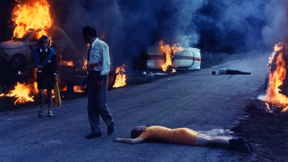 Corinne and Roland stand among burning cars as a woman lies stretched across the road