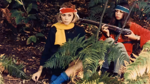 Corinne and a woman with a rifle sit among ferns in the woods