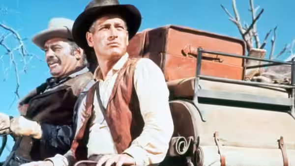 John Russell (Paul Newman) rides beside a mustached man on a wagon