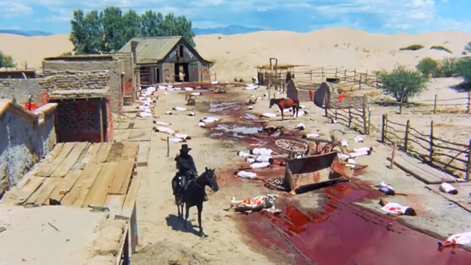A lone rider approaches a dusty settlement strewn with bodies and blood