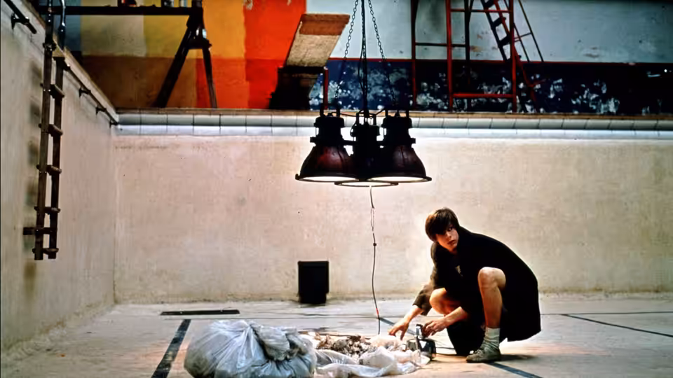 A young man kneels beside a person lying beneath a hanging lamp in an indoor pool