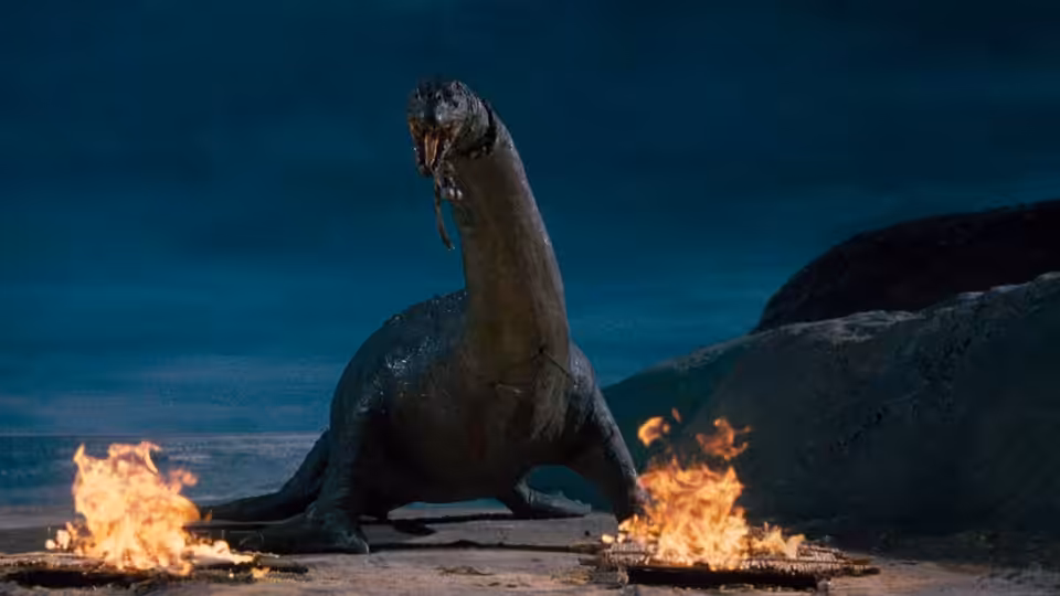 A long-necked dinosaur stands beside flames on a rocky shore
