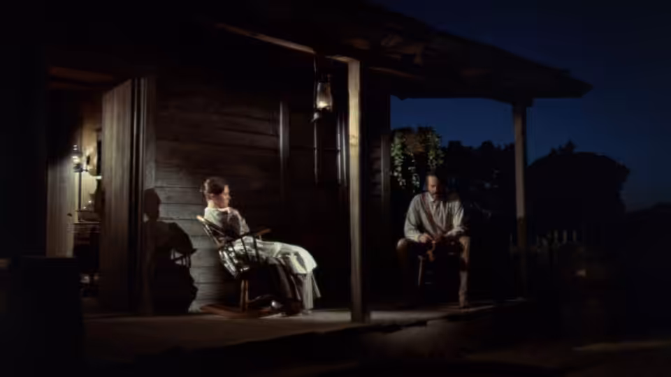 A woman sits on a porch while a man watches from the darkness outside