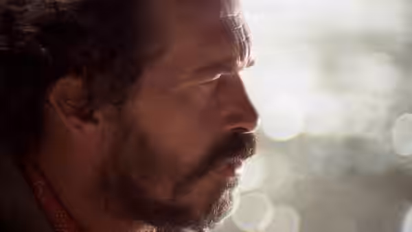 A bearded man looks downward in profile against a bright, blurred background