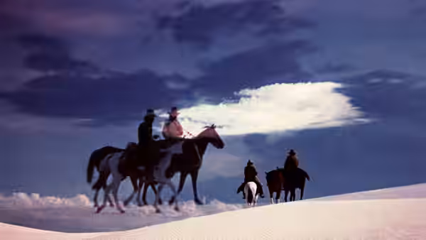 Four riders cross a pale ridge beneath a vast evening sky