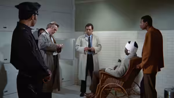 A man in a brown jacket stands beside a seated patient as doctors and a police officer watch