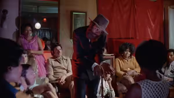 A man in a fedora leans toward a child seated among a gathering of people
