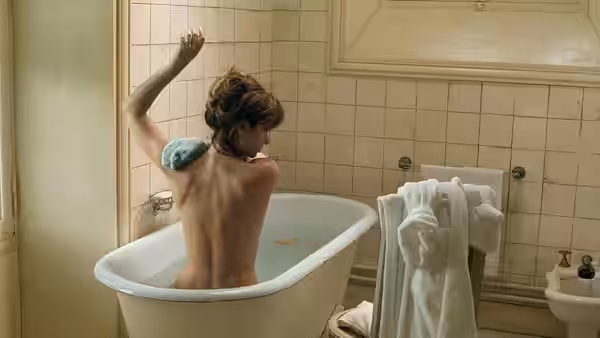 A woman sits naked in a bathtub, reaching toward the tiled wall