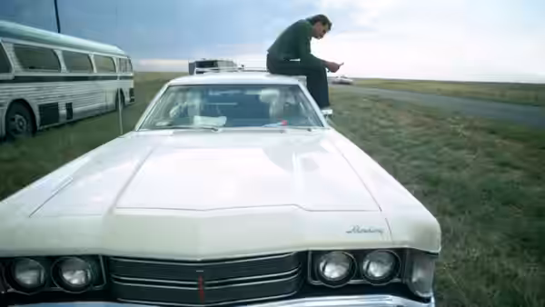 A man stands atop a white car beside a bus on a flat roadside