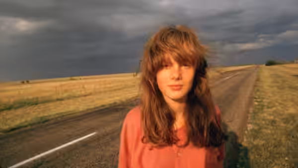 The Girl (Laurie Bird) stands on a straight road through open fields