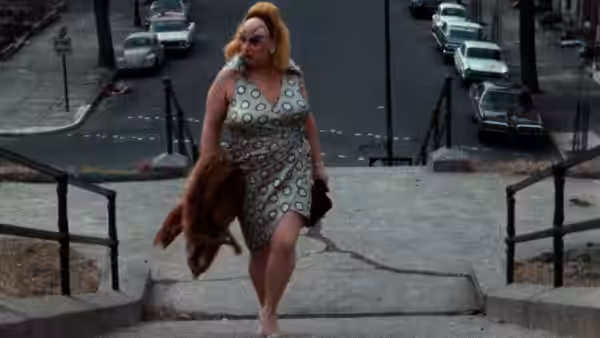 Divine (Divine) descends outdoor steps in a patterned dress, carrying a brown handbag