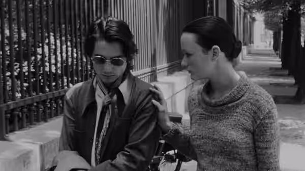 Alexandre (Jean-Pierre Léaud) in sunglasses sits beside a woman in a courtyard