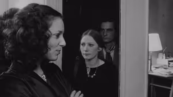 A curly-haired woman faces another woman as a man watches from the doorway