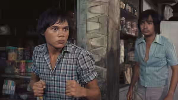 A young man clutches his shirt in a shop while another man watches from the doorway