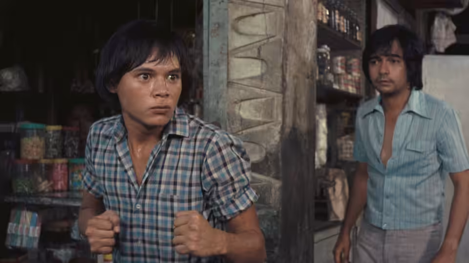 A young man clutches his shirt in a shop while another man watches from the doorway