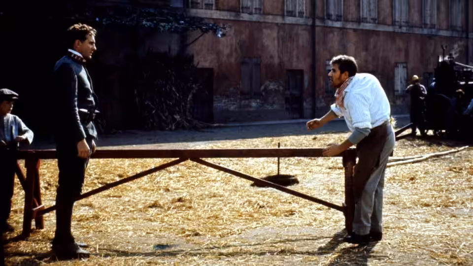 A suited young man faces a worker across a wooden threshing frame in a straw-covered courtyard