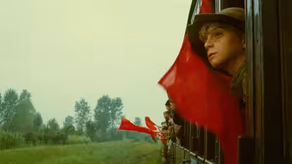 A young passenger leans from a train window beside red flags