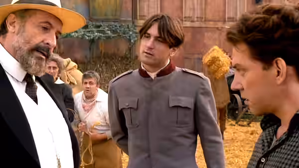 A bearded older man in a hat speaks with two younger men in a farmyard