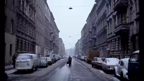 A lone pedestrian walks down a wet city street lined with parked cars
