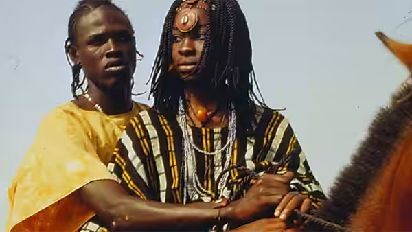A young man in beaded braids sits beside another man near a horse