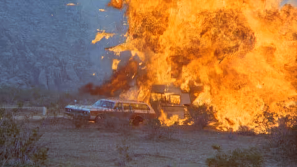 A station wagon burns beside a desert road as flames engulf the vehicle