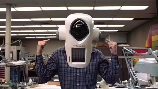 A man in a plaid shirt wears a large white robot helmet in a laboratory