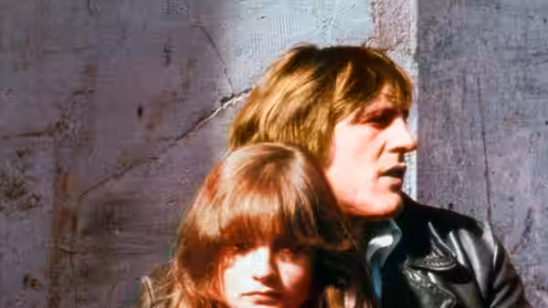 Loulou (Gérard Depardieu) stands close behind Nelly (Isabelle Huppert) beside a stone wall