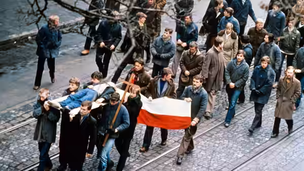 A crowd marches along a cobbled street carrying a Polish flag