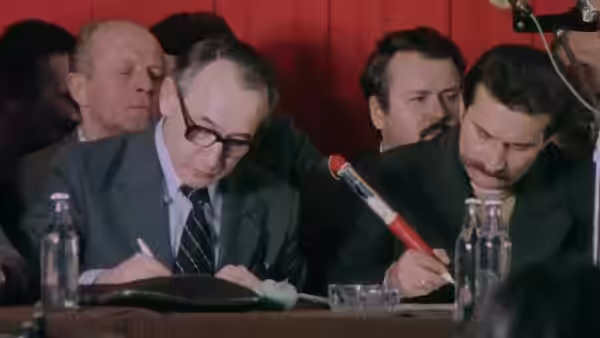 A man writes at a conference table as officials sit beside him
