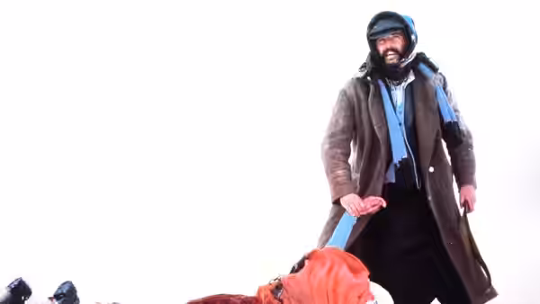A bearded man in a long coat stands over a person lying in the snow