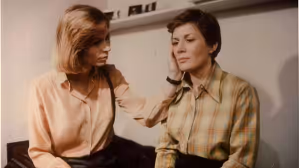 A seated woman gently touches another woman's cheek in a plain room