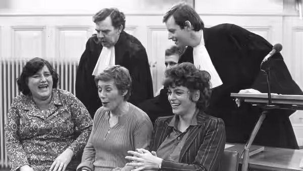 Three women sit together in court as two robed lawyers stand behind them