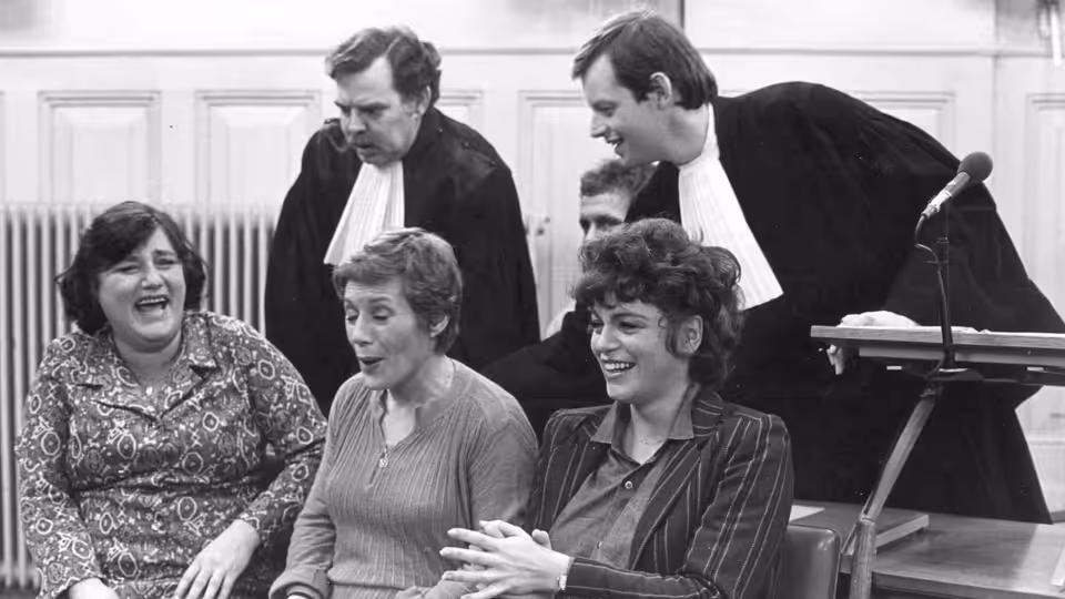 Three women sit together in court as two robed lawyers stand behind them