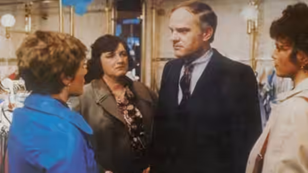 Three women surround a suited man inside a brightly lit room