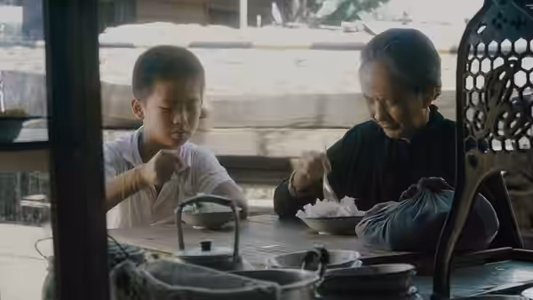 A boy eats at a table beside an elderly woman in a traditional home