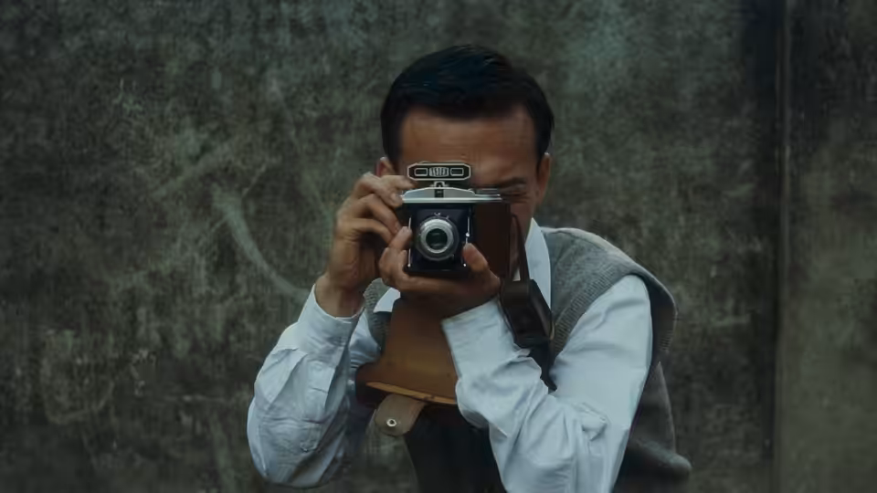 A man raises a camera to his eye against a weathered wall